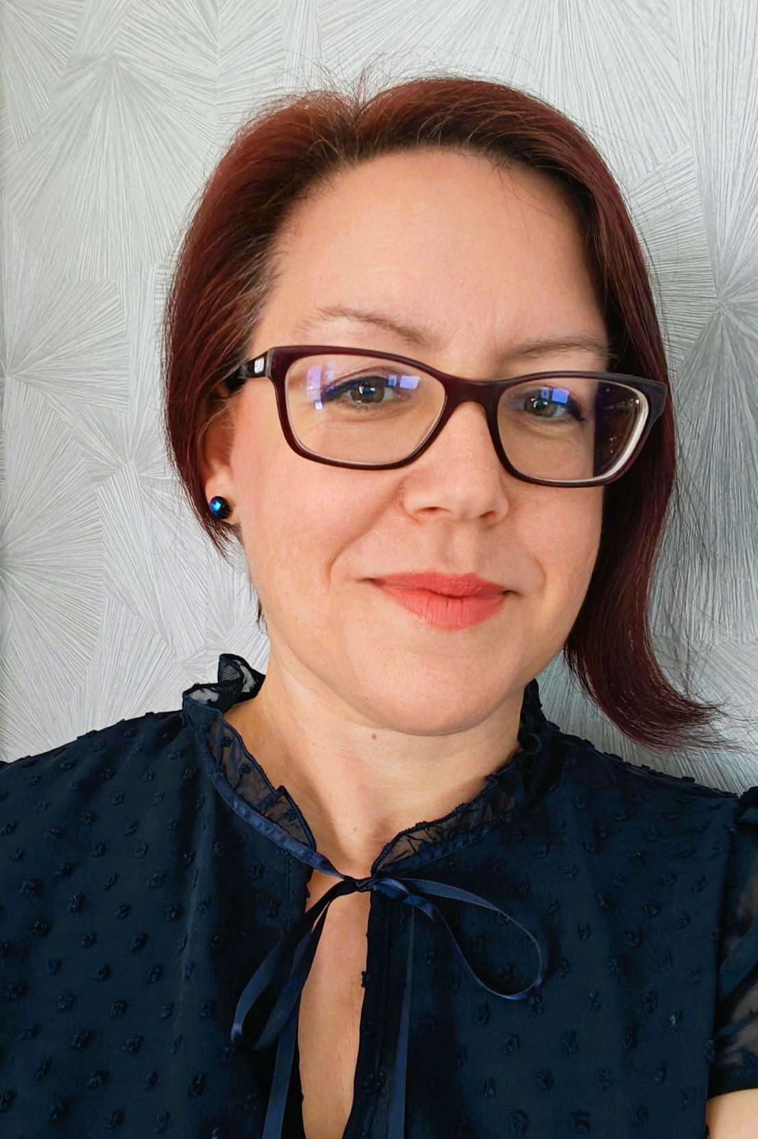 Vörös hajú nő bordó szemüveggel, kék mintás blúzban, kék fülbevalóval, semleges háttér előtt.